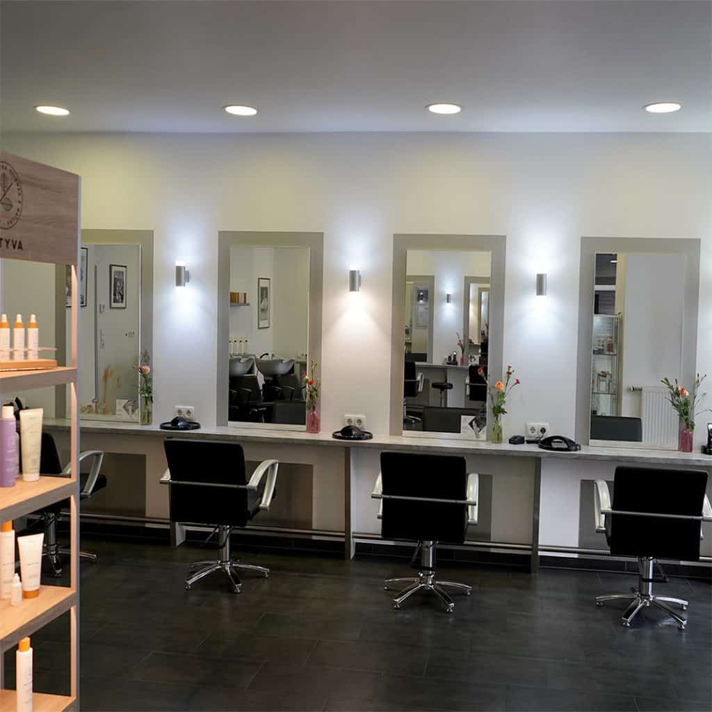 Friseursalon Velly Coiffure in Koeln-Ehrenfeld von innen. Zu sehen sind Frisiertische mit Stühlen und Spiegeln und an der linken Seite ein brauner Holzregal mit Pflegeprodukten.
