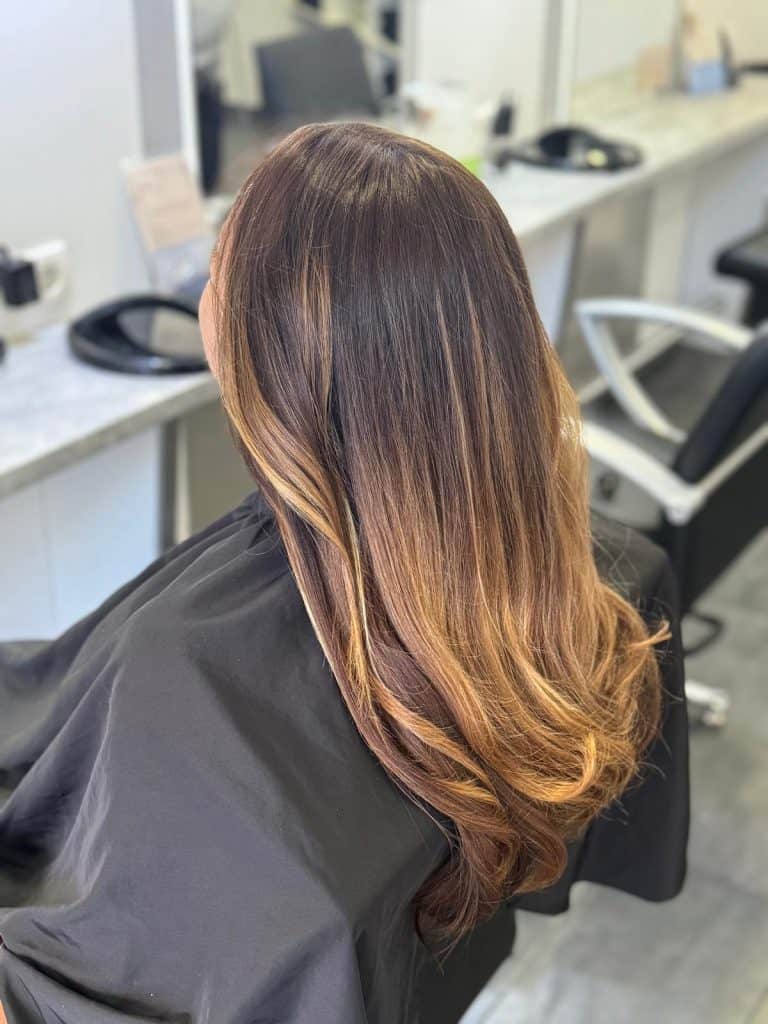weibliche Kundin im Salon, auf einem Frisierstuhl sitzend vor dem Spiegel, von hinten zu sehen, mit langen welligen braunem Haar mit heller werdenden Tönen an den Enden.
